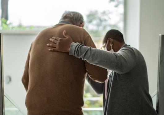 Rear view photo of health care worker helping a patient to walk, inside a room with windows