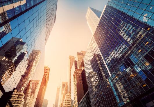 Photograph of tall office buildings, taken from the street, looking up to a sunny sky and tops of skyscrapers