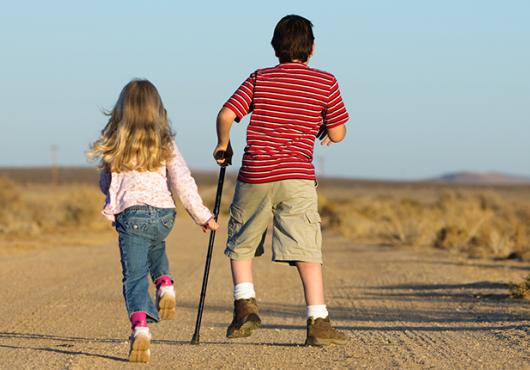 Two children - one girl unassisted and one boy with a crutch - skip down a dirt road