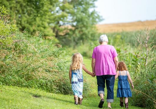 Woman in pink top walking, holding hands with two little girls, backs to camera, in a garden