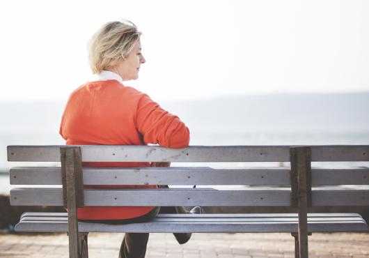 Woman with light hair and orange sweater sits alone on a bench overlooking water