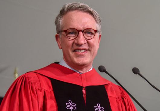 A white man in glasses and red robes smiles at a microphone