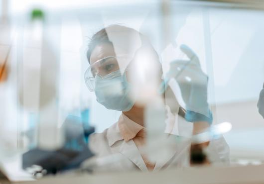 A woman in safety glasses, mask and lab coat working at a laboratory table seen through the reflections and refractions on a safety glass window.