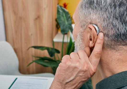 back of the head of a white grey-haired man holding a finger to his ear that has a hearing aid