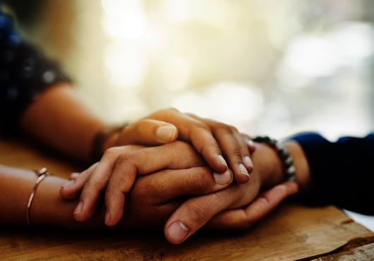 Closeup of two people’s hands clasped together on a table in a comforting manner.