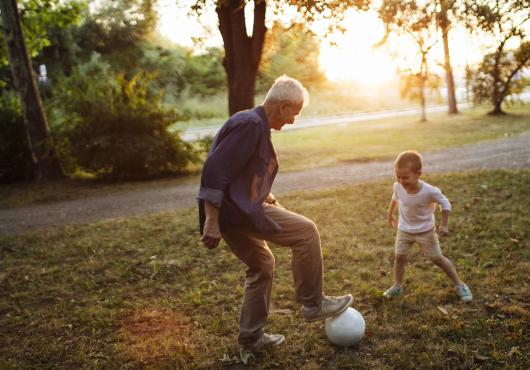 Caught in the middle of a one-on-one soccer game, a wite-haired man stands with his foot on a soccer ball, a small child faces him, poised to take the ball.