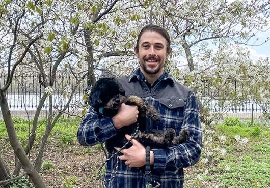 Nelson LaMarche with his miniature poodle in a NY city park with blooming pear tree and Hudson River behind him