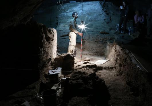 Man with lamp stands in a cave, an excavated skull in front of him