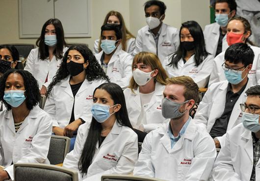 17 students wearing masks and in white coats that say Harvard Medical School seated in auditorium seats
