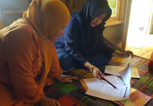 Two women, one in orange and one in blue seated on a colorful rug look at documents together.