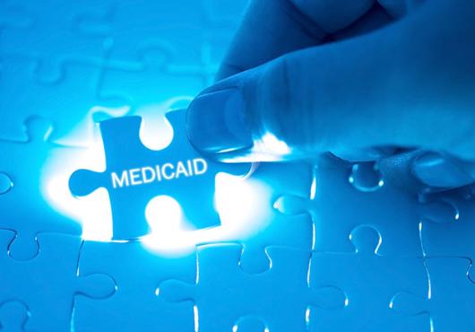 A hand fitting a puzzle piece adorned with the word "medicaid" into a back-lit jigsaw puzzle.