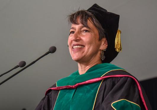 Close-up of a white woman with gray hair in doctoral robes smiling as she speaks into a microphone
