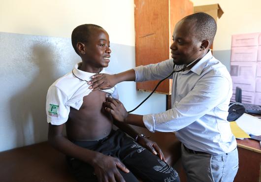 A teenage boy sits on an examination table in a clinic as a man listens to his heart with a stethoscope.