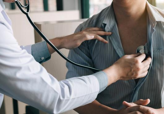 A person in a white jacket is using a stethoscope to listen to the heartbeat of person with a loosely buttoned striped shirt.