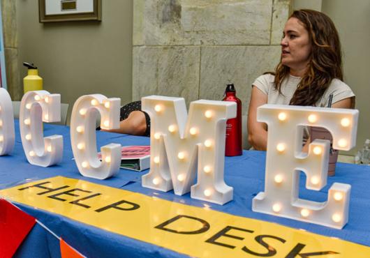 A women sits behind a table with lit up signage letters the spell out OCCME