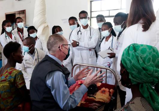 A line of women and men wearing white lab coats, medical masks, and stethoscopes stand facing a three people, including Paul Farmer, sitting on hospital beds. 