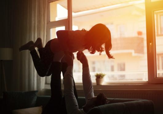 A father plays with his daughter backlit by sunset, seen through the window.