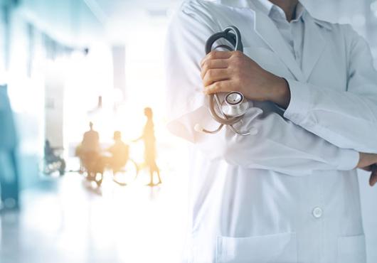 A physician with arms crossed stands in front of a blurry, overexposed background of a mostly empty waiting room.