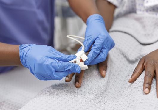 Close up shot of a medical professional placing a pulse oximeter on the finger of a Black hospitalized patient who is lying in bed.