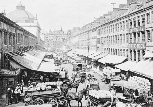An antique black and white photo of the carts and stalls at Boston's Quincy Market from the 19th century. 