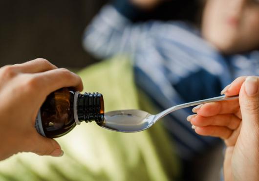 Closeup of adult women's hands pouring cough medicine into a spoon with child blurred out in background