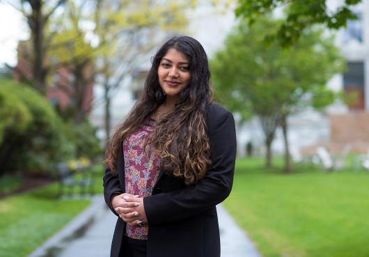 A woman with long, curly hair wearing a dark blazer and a flower-patterned shirt stands on a path between spring green bushes and lawn, an out of focus marble building in the background.