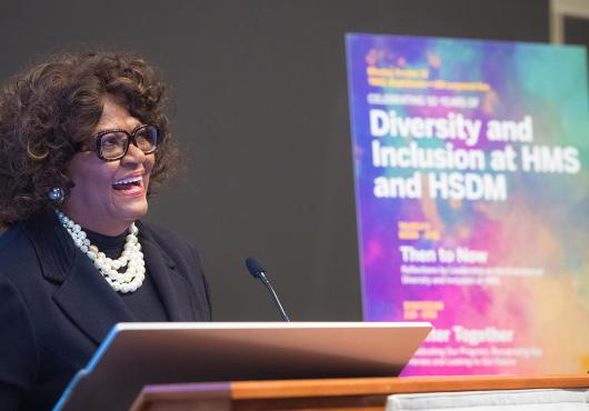 Alt Text:  A woman standing at a lectern, speaking in front of a colorful, out-of-focus poster