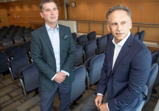 Two men in suits lean against chairs in an auditorium. 