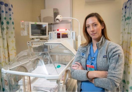 A woman in scrubs and a fleece stands with arms crossed next to a neonatal cradle.