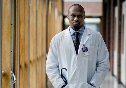 Stultz in a white coat with stethoscopes and hands in his pockets standing in a hallway