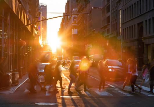 A group of people cross a street at a crosswalk, backlit by the glow of the sun and surrounded by a halo of rainbowed lens flare.