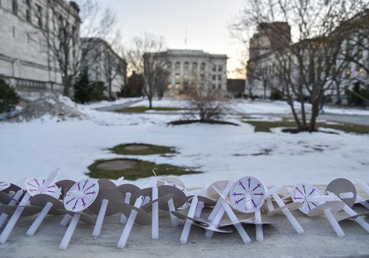 Unlit candles and paper holders line a stone wall at the edge of a snowy HMS Quadrangle, with Gordon Hall visible in background