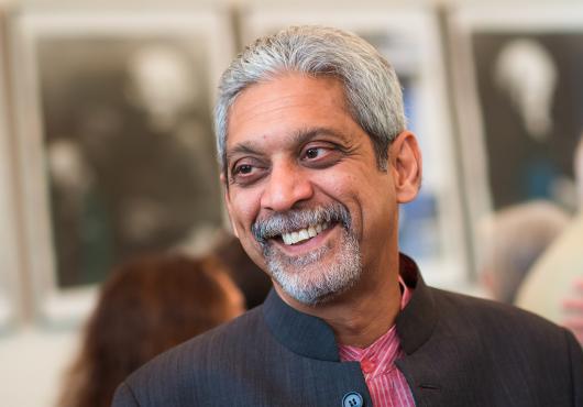 A silver-haired man, wearing a black jacket and a red shirt, smiles at someone off camera.