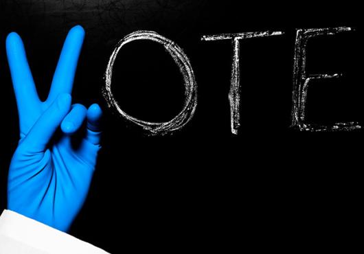 Photo of chalkboard with the word vote - the V is being made by the hand of a person with surgical gloves on.