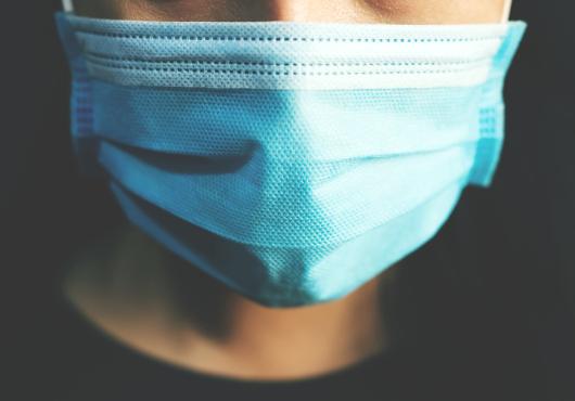 Closeup of the face of a person wearing a paper medical mask in front of a dark background.