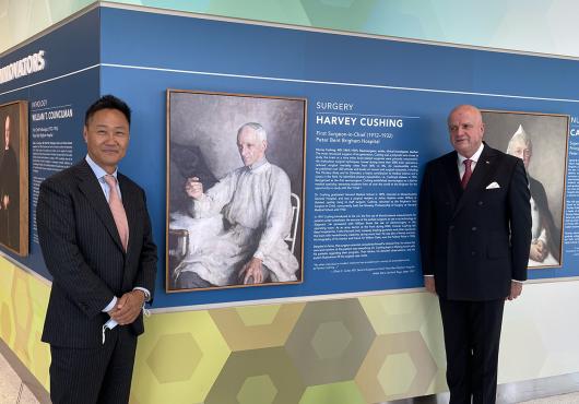 In a hallway painted with colorful geometric patterns, two men in suits stand on either side of a portrait of a man in surgical gowns reviewing an X-ray of a patient’s skull. The portrait is labeled “Harvey Cushing.”