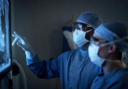 Two figures wearing surgical masks, gowns, and caps examine a backlit X-ray image