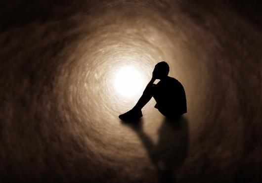 The silhouette of a young person in shorts and t-shirt covering their face as they sit in a dark tunnel.