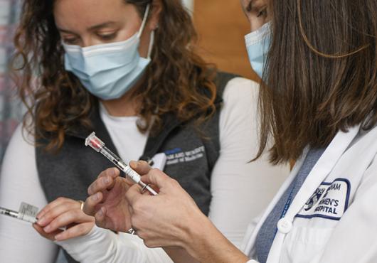 Two women in white coats and surgical masks hold syringes labeled "COVID-19 vaccine"