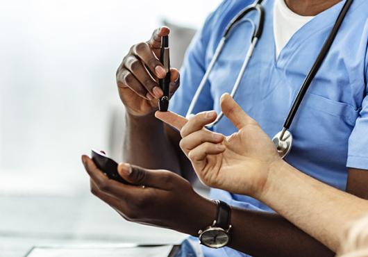 Health care provider in blue scrubs with stethoscope and darker skin tests the blood sugar of a light-skinned patient 