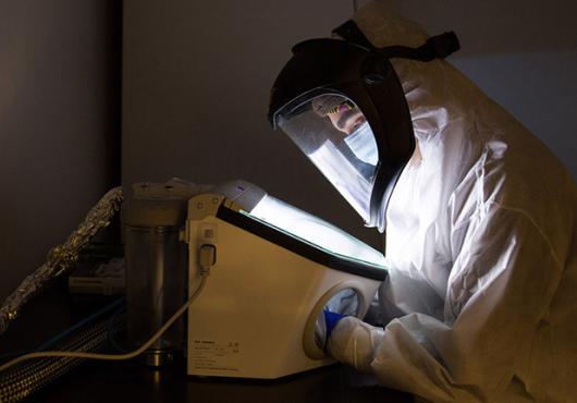 Person in a full-body clean suit and masked hood leans over a lit display in a dark room