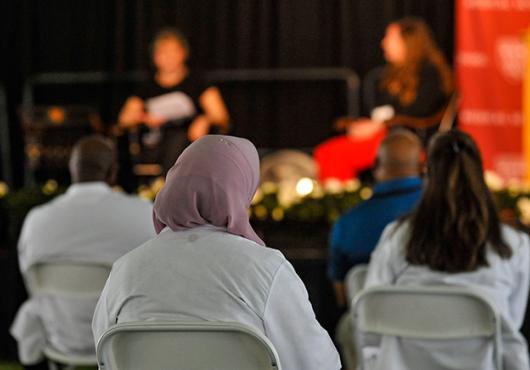 Three people in white coats in focus in audience watch a female patient and female doctor on stage out of focus