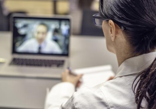 Looking over a doctor's shoulder, we see a patient video conferencing on the doctor's laptop.