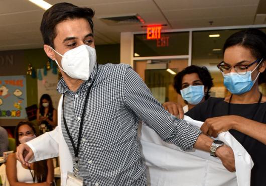 Male student in check shirt wearing a mask puts on a white coat with help of woman