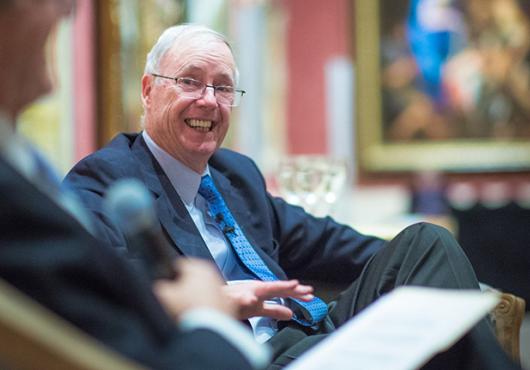 Candid photo of a gray-haired man in glasses grinning in a chair while being interviewed