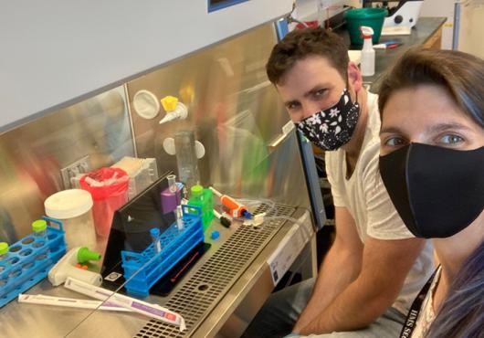 Two people in masks smile beside a hooded lab enclosure