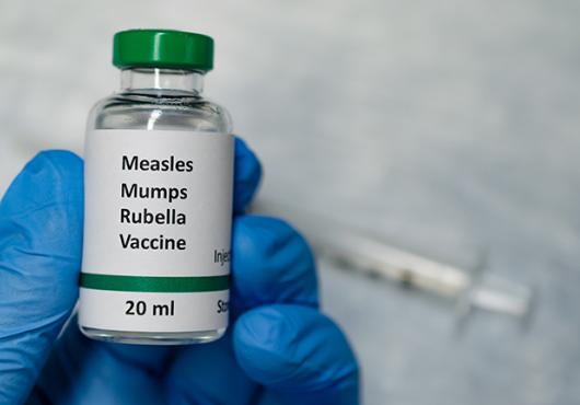 Gloved hand holds a vial labeled "Measles mumps rubella vaccine 20 milliliters" with syringe in background