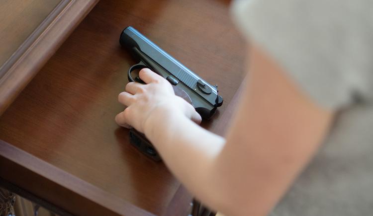 A child's hand reaches into an open drawer in a wooden dresser to pick up a black handgun with a brown handle.
