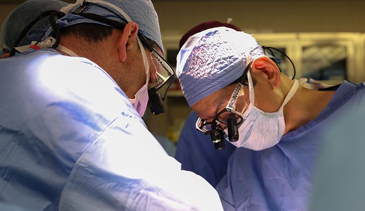 Photo of surgeons in scrubs and masks operating on an unseen patient. Only one is seen head-on. He wears attachments on his glasses to magnify what he's seeing.
