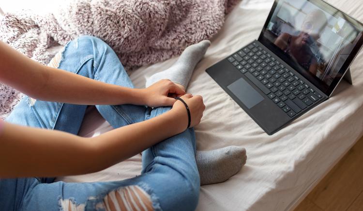 Her face offscreen, a young woman sits on a couch with her arms and legs crossed, wearing ripped blue jeans. At her feet, a laptop shows a face, the details obscured by sun glare. A telemedicine therapy session is underway.